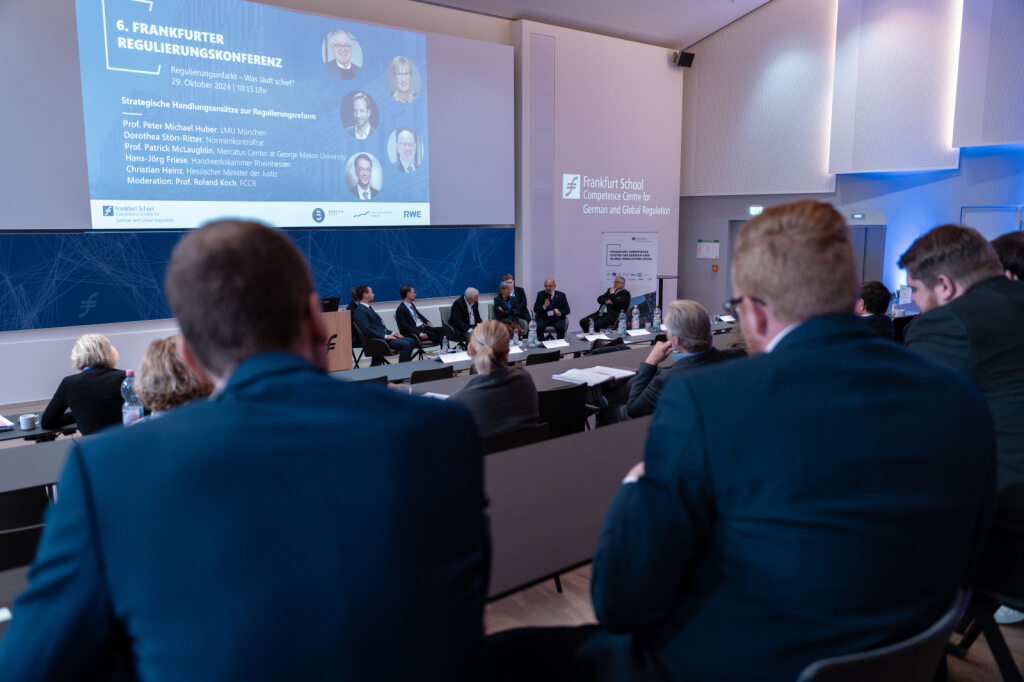 Eine Podiumsdiskussion mit mehreren Personen auf einer Bühne. Im Vordergrund sitzen Zuhörer in einem Hörsaal. Auf der Leinwand steht „6. Frankfurter Regulierungskonferenz“.